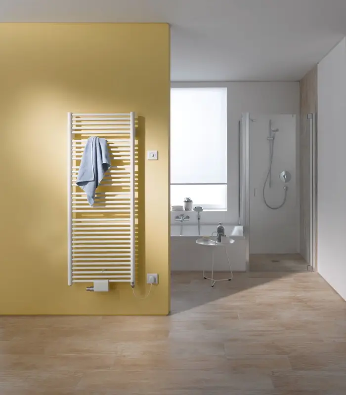 Vertikaler weißer Kermi-Heizkörper mit schmalen Längsrippen, an einer gelben Wand montiert, neben einem modernen Badezimmer mit Dusche und Badewanne.